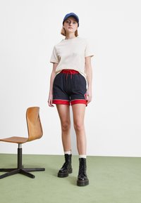 Camiseta beige, pantalones cortos azul marino con ribete rojo y cordón, combinados con botas negras de cordones. Silla de madera y alfombra verde en el fondo.