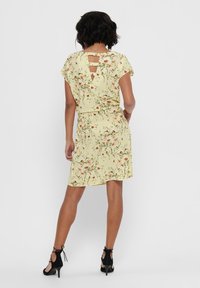 Robe jaune à manches courtes avec des motifs floraux, un décolleté en trou de serrure dans le dos, et une taille cintrée. Portée avec des talons ouverts noirs.