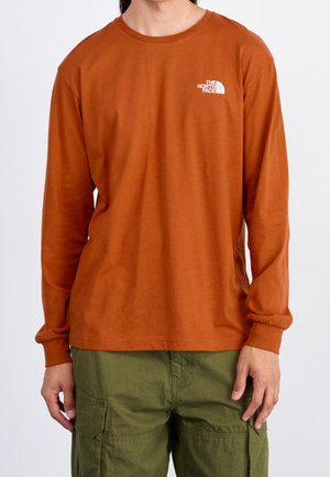 Mann trägt ein langärmliges Hemd in verbranntem Orange mit dem The North Face-Logo auf der Brust und olivgrüne Cargohosen.
