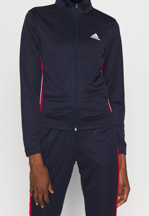 Close-up of person wearing black Adidas zip-up jacket and matching pants with red stripes on sides, hands relaxed by sides.