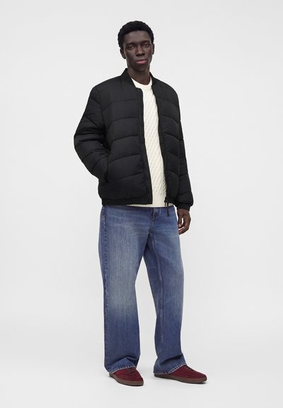 Jeune homme portant une veste matelassée noire sur un pull en maille blanc, un jean bleu et des baskets rouges, debout avec une main dans la poche.