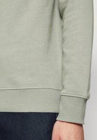 Lichtgroene trui met een gestructureerde afwerking, geribde manchetten en een afgeronde zoom; gecombineerd met donkerblauwe spijkerbroek.