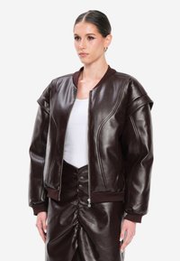 Giacca bomber in pelle marrone con polsini e scollo a coste, caratterizzata da una zip frontale e maniche strutturate, abbinata a una gonna coordinata.