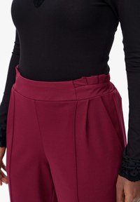 Pantalons taille haute bordeaux avec plis et poches latérales, associés à un haut noir à manches longues avec une accent en dentelle sur les poignets.