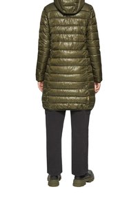 Olijfgroene, gewatteerde lange jas met een capuchon. Gladde, glanzende textuur en horizontale stiksels. Combinatie met zwarte broek en stevige zwarte laarzen.