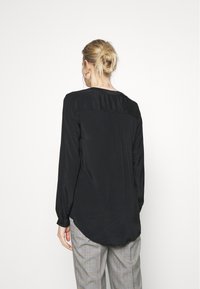 Blusa nera a maniche lunghe con una texture liscia, scollatura rotonda e dettagli plissettati sul retro, abbinata a pantaloni a quadri.