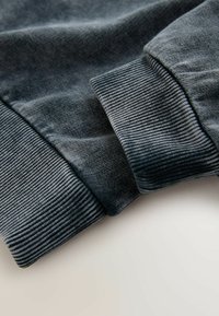 Tumši pelēki džempera aproces ar ribveida tekstūru, mīkstu audumu un gludu apdari. Tuvplāns atklāj šuvju struktūru.