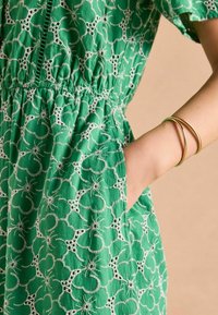 Zelené šaty s bielou kvetinovou výšivkou, elastický pás a vrecká. Modelka predvádza zlaté náramky na zápästí na neutrálnom pozadí.