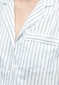 Κοντινό πλάνο ενός ατόμου που φοράει ένα ελαφρώς μπλε και λευκό κάθετα ριγέ πουκάμισο με τσέπη στο στήθος και γιακά με πέτο.