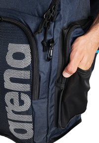 Sac à dos en tissu bleu avec poche en filet, accents noirs et plusieurs compartiments zippés. Présente une main accédant à une poche latérale.