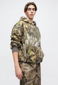 Camo-patterned hoodie featuring earthy tones of brown, green, and tan. Soft fabric with a front pocket and ribbed cuffs. Styled with matching pants.