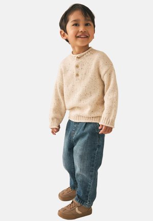 Smilende ung dreng stående iført en cremefarvet strikket sweater med knapper, blå jeans og brune sneakers mod en ensfarvet baggrund.