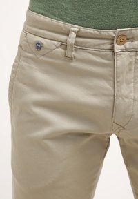 Beige puuvillahousut, joissa on sileä pinta, nappikiinnitys ja pieni sininen logoetumerkki etutaskussa.