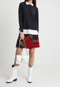 Kvinna iklädd svart tröja över vit skjorta, röd rutiga kjol, vita stövlar, håller en vit handväska med kedjesnodd.