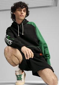 Felpa con cappuccio nera e verde, dotata di tasca frontale e con cordini bianchi, abbinata a pantaloni corti neri e scarpe con dettagli verdi e arancioni.