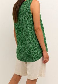 Mujer vestida con una blusa sin mangas verde con pequeños patrones florales negros y pantalones cortos beige hasta la rodilla, de pie frente a un fondo liso.