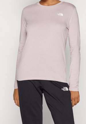 Osoba nosząca jasnoróżową koszulę z długim rękawem i czarne spodnie, obie marki The North Face z logo na klatce piersiowej i udzie.