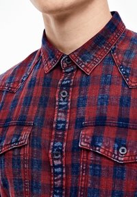 Hedvábně červená a tmavě modrá kostkovaná košile s límcem na knoflíky, dvěma náprsními kapsami a tmavými knoflíky. Materiál vypadá jako texturovaný a tkaný.