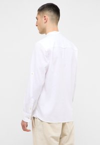 Camicia bianca a maniche lunghe con colletto, dettagli con bottoni sui polsini e un tessuto texturizzato. La vista posteriore mostra una cucitura a yoke e un orlo curvato.