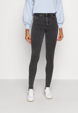 Jeans skinny de cintura alta cinza escuro com um acabamento texturizado, apresentando um fecho frontal com botão e detalhes de costura minimalistas, combinados com ténis brancos.