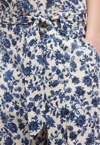 Περιοχή μέσης και γοφών μπεζ παντελονιού με μπλε λουλουδάτο σχέδιο, υφασμάτινη ζώνη δεμένη, κλείσιμο με κουμπί και πλαϊνή τσέπη με χέρι μέσα.