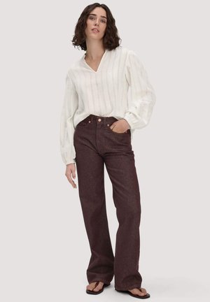 Frau mit mittellangem, lockigem Haar, die eine weiße Bluse mit langen Ärmeln und hoch taillierte, dunkelbraune Jeans mit weitem Bein trägt, mit einer Hand in der Tasche.