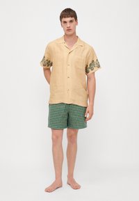 Camicetta beige in lino a maniche corte con dettaglio ricamato verde sulle maniche, abbinata a pantaloni corti a quadri verdi e blu navy.