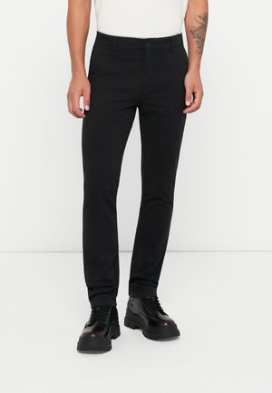 Schwarze Slim-Fit-Hosen aus Baumwollmix, mit sichtbaren Taschen und Gürtelschlaufen. Getragen mit schwarzen Stiefeln mit einer groben Sohle.