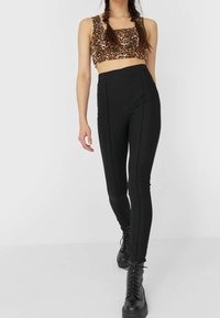 Czarne legginsy high-waisted z widocznymi detalami szwów, zestawione z brązową bluzką w lamparcim wzorze. Modelka nosi czarne sznurowane botki za kostkę.