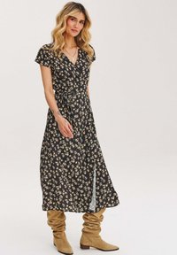 Čierne kvetinové zavinovacie šaty s krátkymi rukávmi, so žltými a bielymi vzormi kvetov, bočným rozparkom a opaskom na viazanie. Hnedé voľné čižmy.