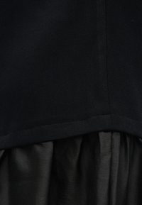 Κοντινή λήψη μαύρου υφάσματος που παρουσιάζει δύο υφές: ομαλή ματ ανώτερη στρώση με ραμμένο τελείωμα και πτυχωτή, ελαφρώς γυαλιστερή κατώτερη στρώση.