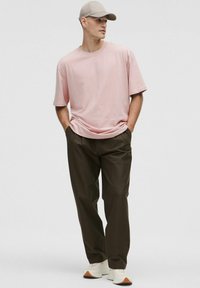 Hombre con camiseta oversize de color rosa claro, pantalones oliva oscuros, zapatillas blancas y gorra beige, de pie con las manos en los bolsillos.