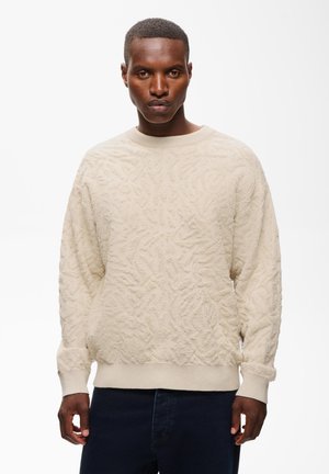 Man met kort haar, dragend een crèmekleurige gemêleerde trui en een donkere broek, staande tegen een effen witte achtergrond.
