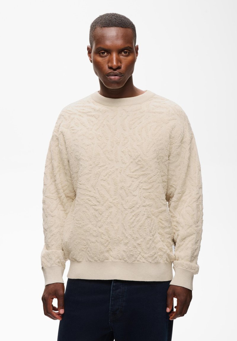 Homme aux cheveux courts portant un pull crème texturé et un pantalon foncé, debout devant un fond blanc uni.