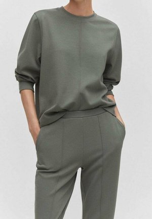 Persona vistiendo un conjunto de manga larga y pantalones color verde oliva a juego, con las manos en los bolsillos, de pie contra un fondo claro y liso.