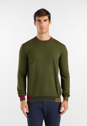 Uomo che indossa un maglione verde oliva a maniche lunghe con strisce colorate su polsini e orlo, abbinato a pantaloni blu scuro, in piedi davanti a uno sfondo bianco.