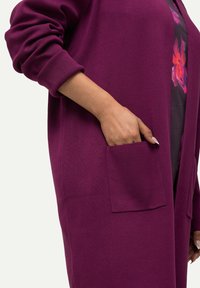 Lila Strickjacke mit langen Ärmeln, gerippten Bündchen und einer vorderen Tasche. Glatte Stofftextur, knielanges Design und lockerer Schnitt.