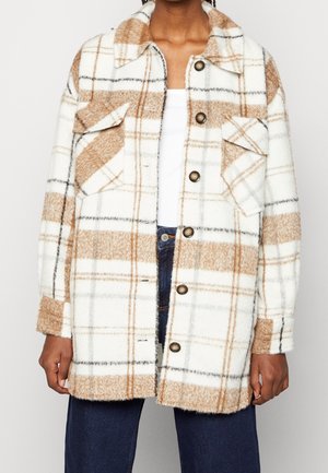 Personne portant une veste à carreaux blanche et beige avec des poches poitrine et un jean bleu foncé, debout devant un fond uni.