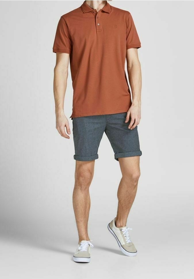 Polo shirt color ruggine con colletto, abbinato a pantaloni corti grigi arrotolati e scarpe da ginnastica leggere. Design semplice, materiale in cotone, senza motivi visibili.