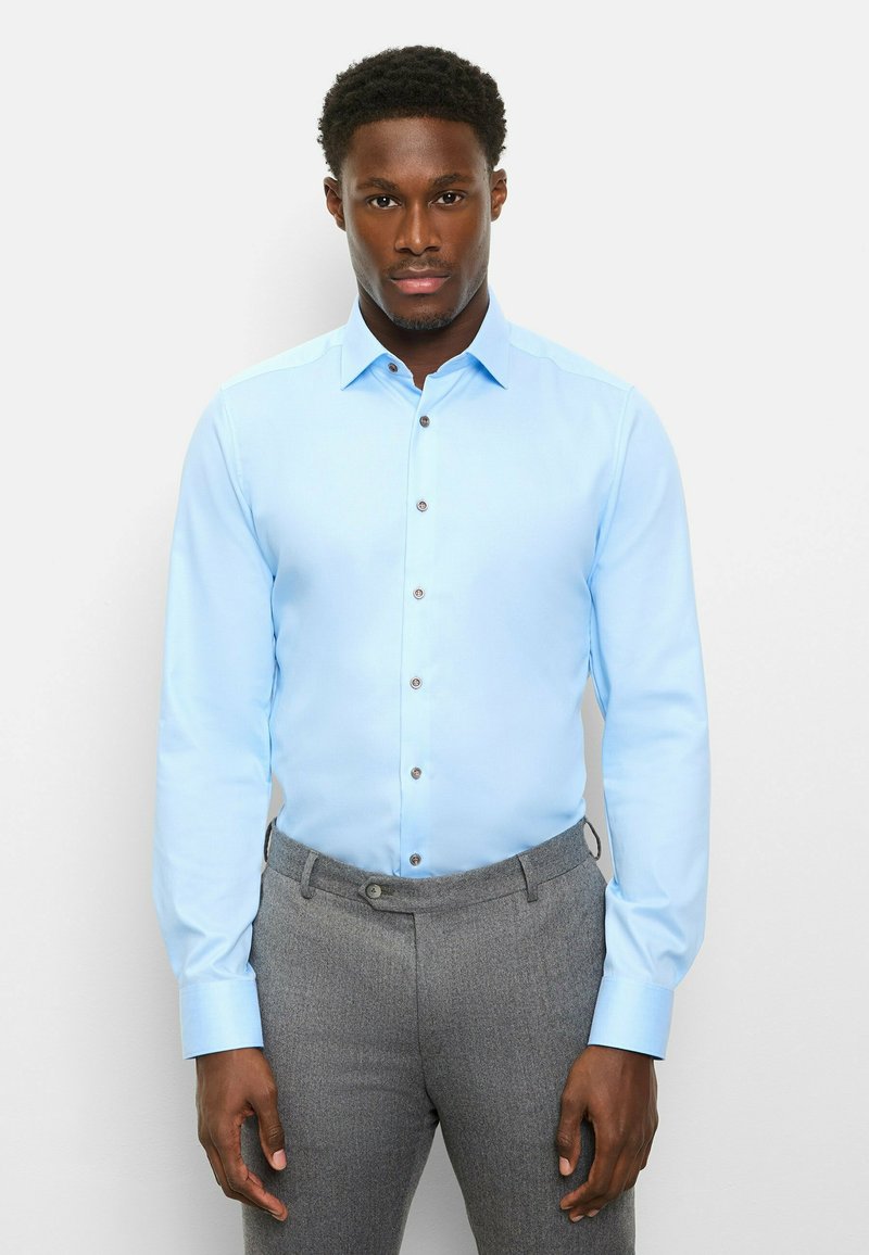 Hellblaues Hemd mit Knöpfen und langen Ärmeln, graue strukturierte Hose und kontrastierende Knöpfe. Glatter Stoff mit maßgeschneiderter Passform.