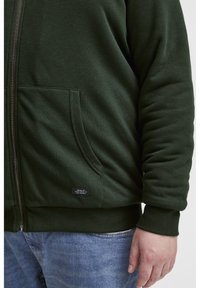 Sudadera verde con cremallera, confeccionada en un tejido suave y con puños acanalados. Presenta un bolsillo frontal y una pequeña etiqueta negra cerca del dobladillo.