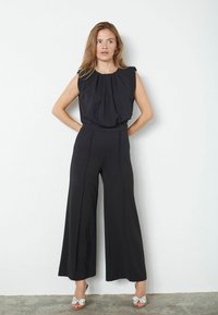 Černý overal s plisovaným vrškem a širokými nohavicemi. Vyznačuje se přiléhavým pasem a hladkým materiálem. Nosený s ozdobnými nazouvacími botami.