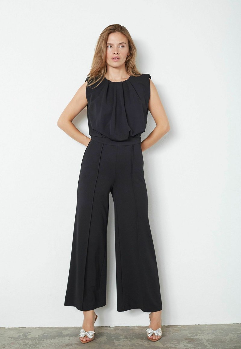 Černý overal s plisovaným vrškem a širokými nohavicemi. Vyznačuje se přiléhavým pasem a hladkým materiálem. Nosený s ozdobnými nazouvacími botami.