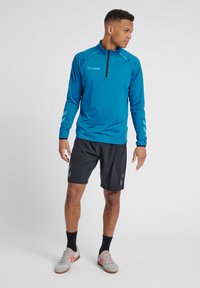 Blå langærmet atletisk top med halv-zip krave, kontrasterende sorte accenter og logo. Kombination med sorte shorts og lysegrå sneakers.