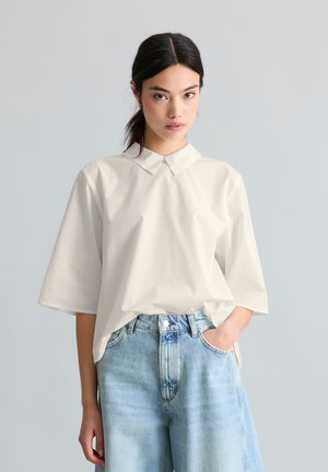 Femme aux cheveux foncés en queue de cheval basse portant une chemise blanche ample à col et un jean bleu clair taille haute à jambes larges, debout avec une main dans la poche.