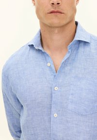 Camicia button-up in lino azzurro chiaro con colletto, presenta una tasca sul petto e un tessuto leggermente testurizzato. Cinque bottoni sul davanti.