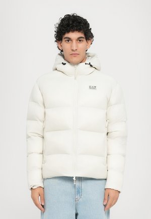 Witte donsjack met capuchon, met een rits aan de voorkant, gevoerde secties en een klein logo op de borst. Draagt over lichtblauwe jeans.