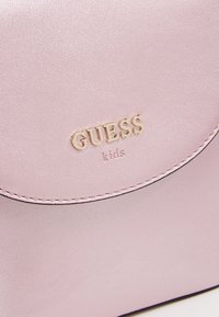 Pieni vaaleanpunainen tekonahkainen laukku, jossa pyöreä läppä, kohopainettu "GUESS kids" -logo kullassa ja ommeltu reunusdetail.