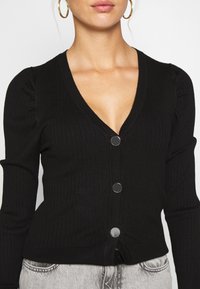 Černý žebrovaný cardigan s hlubokým V-výstřihem, dlouhými rukávy s nabíranými ramenními částmi a kovovými patentkami na přední straně.