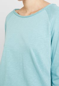 Light blue long-sleeved top featuring a relaxed fit, wide neckline, and textured fabric with frayed edges along the collar.